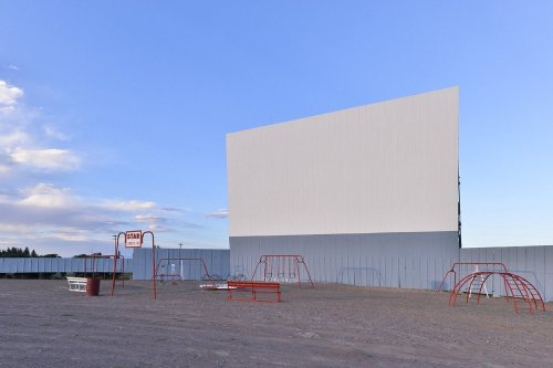 A blank outdoor movie screen stands in an empty lot, surrounded by playground equipment under a clear blue sky.