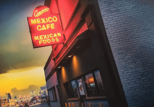 Neon sign for Connie's Mexico Cafe, featuring "Mexican Foods," against a colorful sunset backdrop.