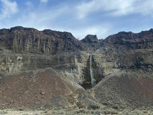 A waterfall cascades down a rocky cliff, surrounded by arid terrain and a blue sky with scattered clouds.