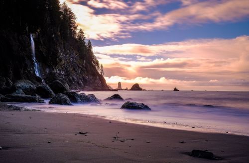 A serene beach scene at sunset, featuring a waterfall, rocky shoreline, and soft waves under a colorful sky.