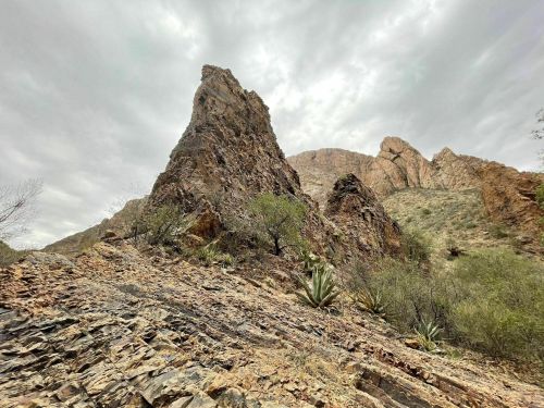 A rugged rock formation rises amidst a desert landscape with sparse vegetation under a cloudy sky.