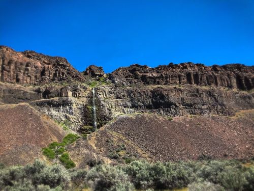 A rocky cliff with a small waterfall cascading down, surrounded by dry vegetation under a clear blue sky.