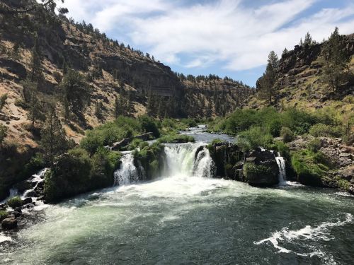 A scenic view of a waterfall cascading into a river, surrounded by lush greenery and rocky cliffs under a blue sky.