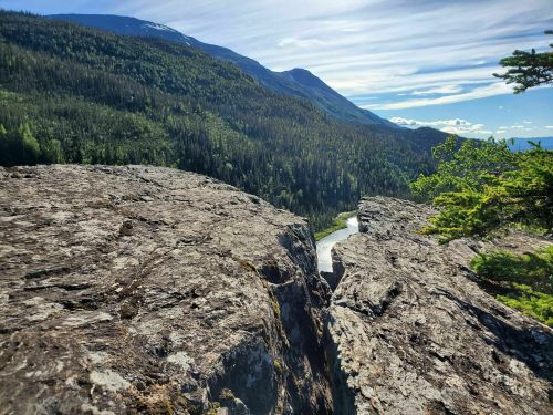 A rocky cliff overlooks a winding river surrounded by lush green forests and mountains under a clear blue sky.