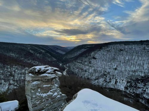 A snowy landscape with a rocky outcrop overlooking a valley at sunset, surrounded by hills and a cloudy sky.
