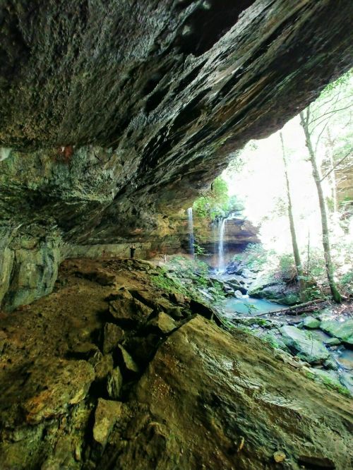 A rocky cave interior with a waterfall in the background, surrounded by lush greenery and sunlight filtering through trees.