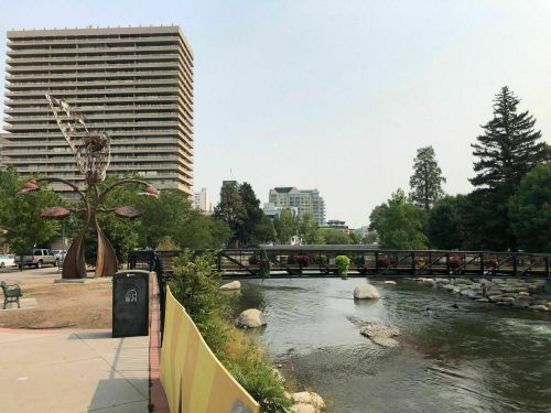 A serene park scene featuring a river, a modern sculpture, and buildings in the background under a clear sky.