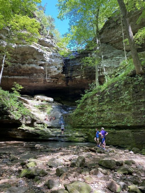 A serene natural scene featuring a waterfall cascading down rocky cliffs, with people exploring the lush surroundings.