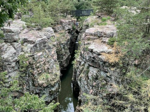 A narrow canyon with rocky cliffs and a small stream running through, surrounded by trees and a wooden bridge above.