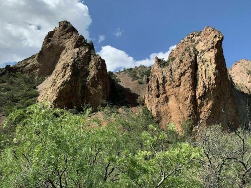 Two rugged rock formations rise against a blue sky with scattered clouds, surrounded by green vegetation.