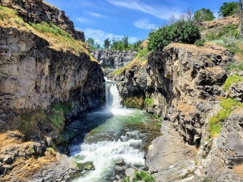 A scenic view of a waterfall cascading through rocky cliffs, surrounded by greenery and a clear blue sky.