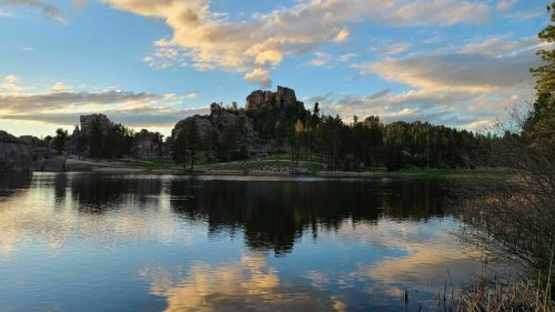 A serene lake reflects rocky hills and a colorful sky with clouds, surrounded by trees in a tranquil landscape.