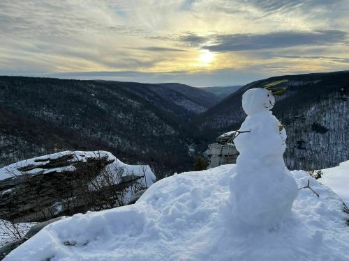 A snowman stands on a snowy ledge, overlooking a valley at sunset with a colorful sky and distant mountains.