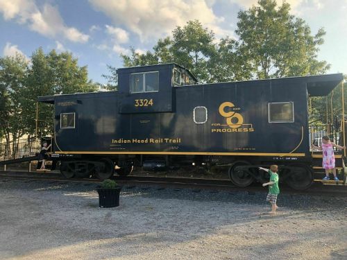 A blue caboose labeled "3324" on a rail trail, with children playing nearby and trees in the background.