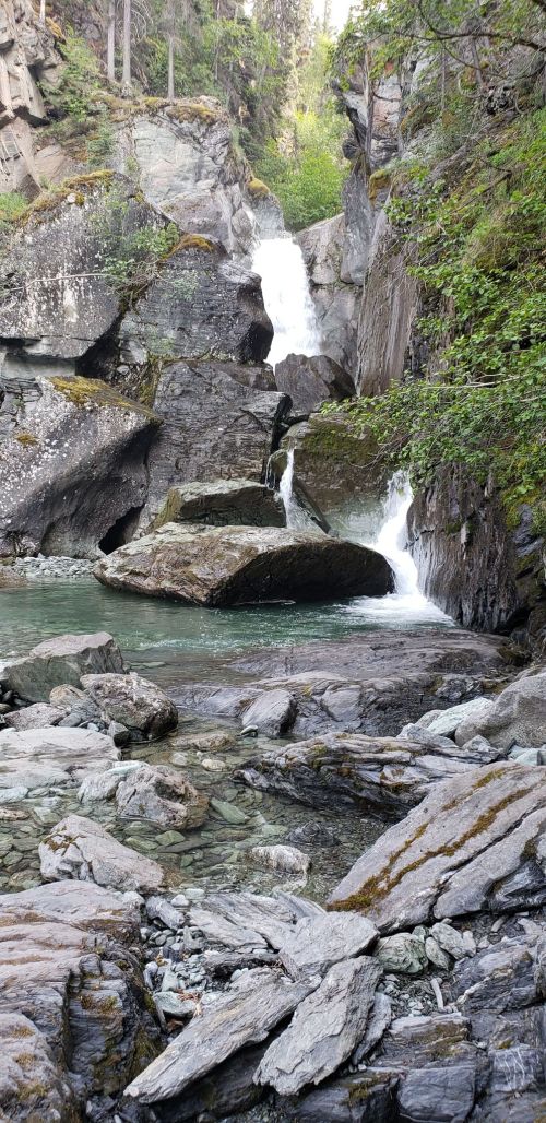 A serene waterfall cascades over rocky cliffs into a clear pool, surrounded by lush greenery and smooth stones.