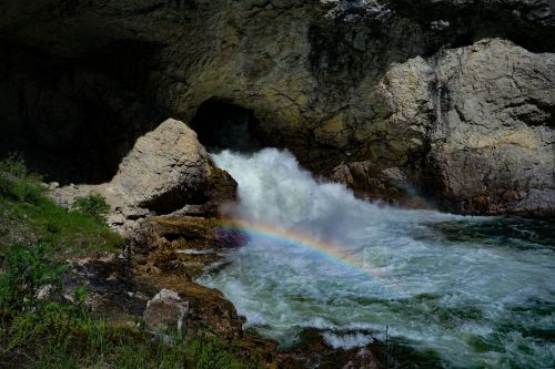 A waterfall cascades over rocks, creating a rainbow in the mist against a rugged, rocky backdrop.