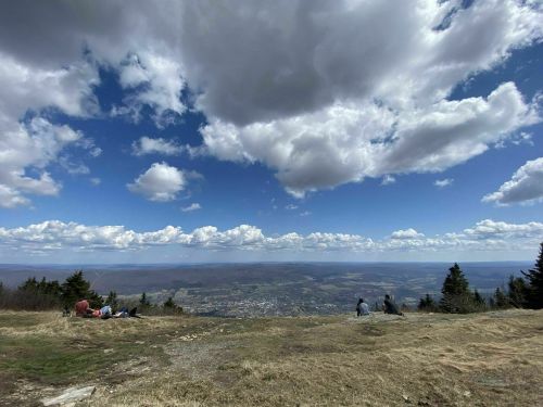 A panoramic view from a mountain top, featuring a blue sky with clouds and a valley below. People are visible in the foreground.
