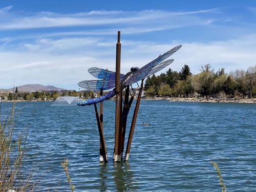 A colorful dragonfly sculpture stands on metal poles in a serene lake, surrounded by trees and a clear blue sky.