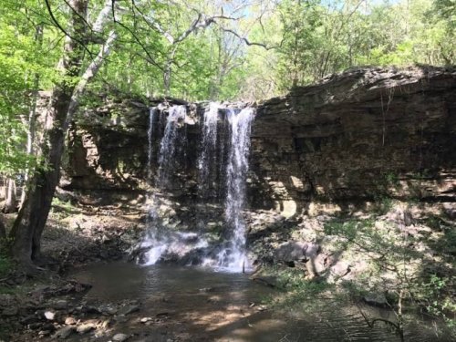 A serene waterfall cascading over rocky cliffs into a calm stream, surrounded by lush green trees.