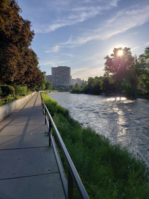 A serene riverside path lined with trees, with a city skyline in the background and the sun setting over the water.