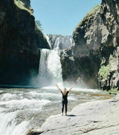 A person stands with arms raised in front of a majestic waterfall, surrounded by rocky cliffs and greenery.