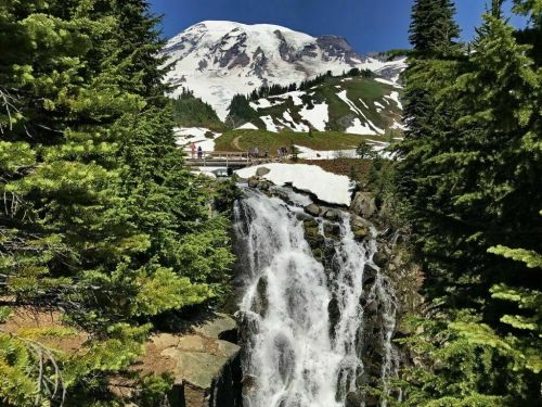 A waterfall cascades down rocky terrain, surrounded by lush greenery and snow-capped mountains in the background.