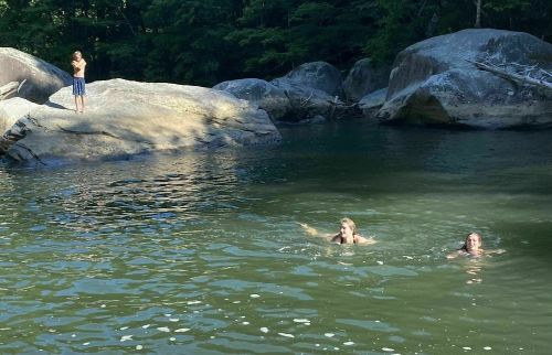 Two people swim in a calm river surrounded by large rocks, while a third person stands on a rock nearby.