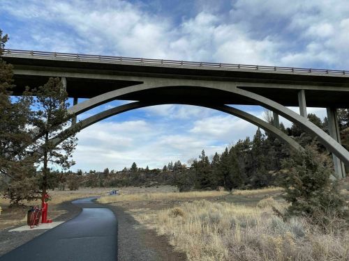 A concrete bridge arches over a path surrounded by dry grass and trees under a cloudy sky.