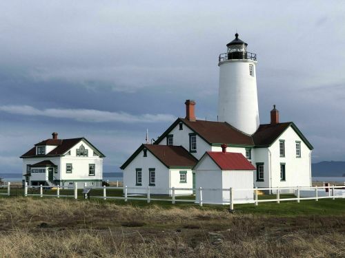 A white lighthouse with a red roof, accompanied by a nearby white house, set against a cloudy sky.