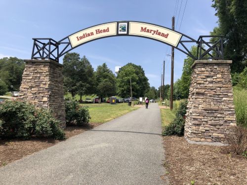 Archway sign reading "Indian Head Maryland" over a pathway lined with greenery and trees.