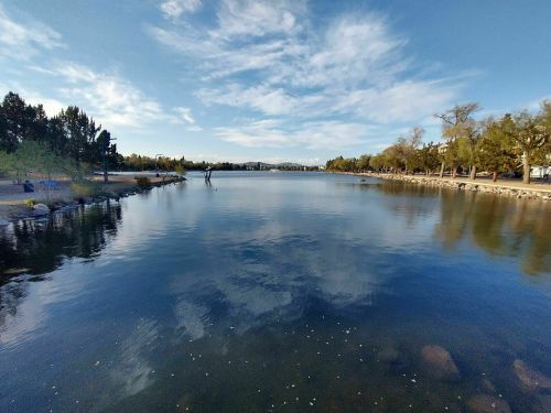 A serene lake reflecting the sky, surrounded by trees and a walking path, with a person fishing in the distance.