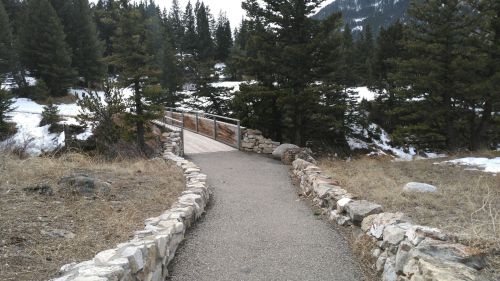 A winding stone path leads to a wooden bridge surrounded by trees and snow-capped mountains in the background.