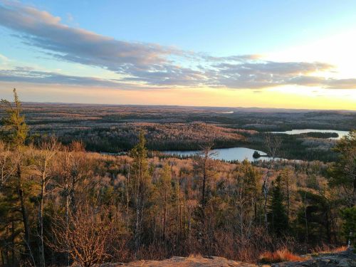 A scenic view of a forested landscape at sunset, featuring lakes and rolling hills under a colorful sky.