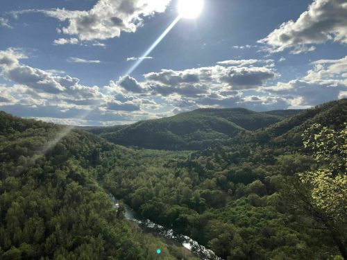 A scenic view of lush green hills and a winding river under a bright sun and partly cloudy sky.