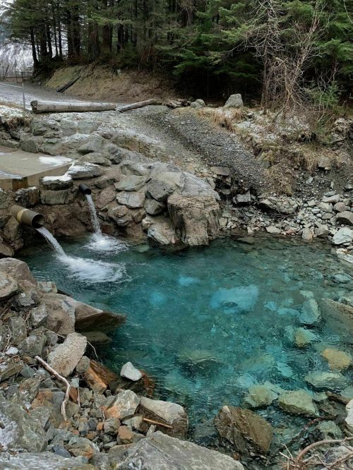 A clear blue pool surrounded by rocks and trees, with water flowing from a pipe and small waterfalls nearby.