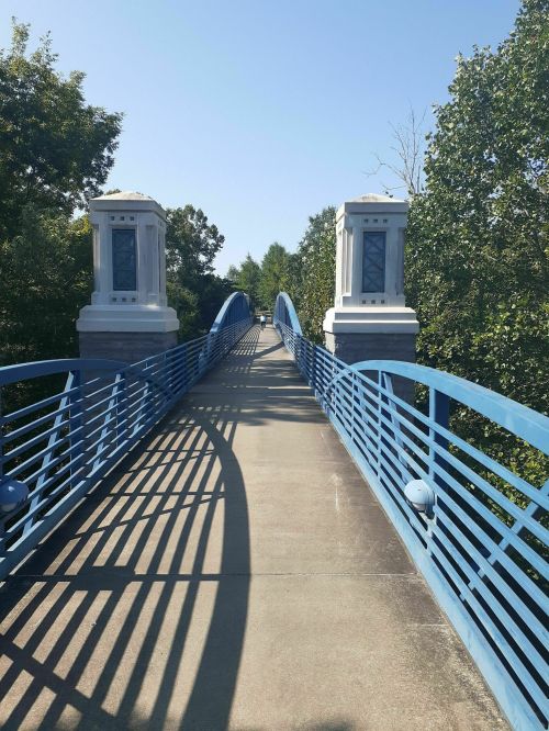 A blue pedestrian bridge with decorative pillars, surrounded by trees under a clear blue sky.