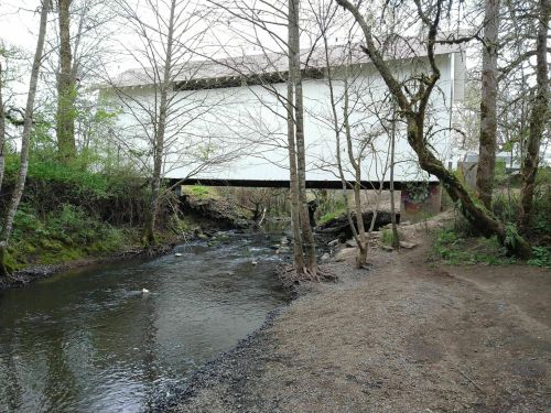 A covered bridge spans a small creek, surrounded by trees and a gravel path along the water's edge.