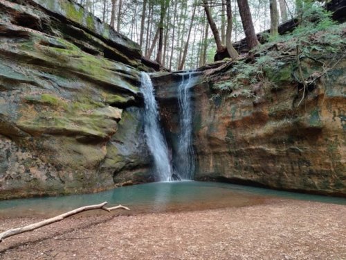A serene waterfall cascades into a clear pool, surrounded by mossy rocks and lush green trees.