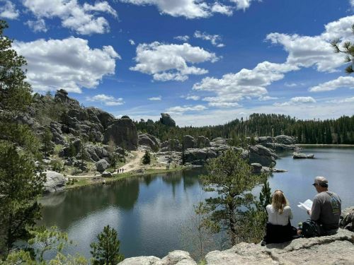 A serene landscape featuring a lake surrounded by rocky formations and trees, with two people sitting on a ledge.