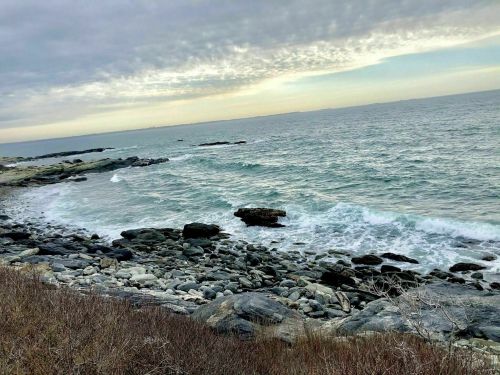 A rocky shoreline with gentle waves lapping against the stones under a cloudy sky.