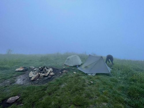 A foggy campsite with two tents and a fire pit surrounded by grass and rocks.