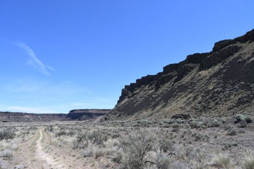 A wide, arid landscape with a dirt path leading towards rocky cliffs under a clear blue sky.