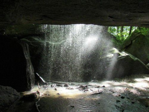 A serene waterfall cascading through a rocky opening, with sunlight creating a misty effect in a lush green setting.