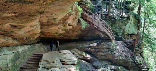 Two hikers explore a rocky area with a wooden bridge and lush greenery in a forested landscape.