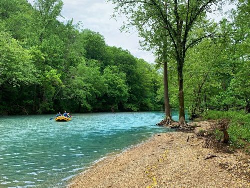 A yellow raft with people floats on a clear blue river, surrounded by lush green trees and a sandy shore.