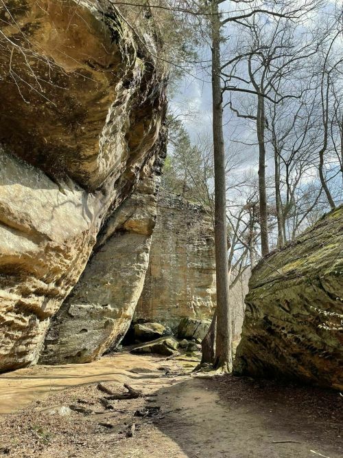A narrow path between towering rock formations and trees under a cloudy sky.