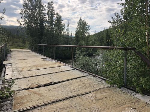 A weathered wooden bridge surrounded by lush greenery and trees, with a river visible in the background.