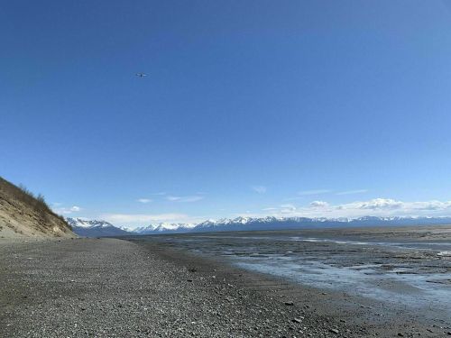 A serene beach scene with a clear blue sky, mountains in the distance, and a small plane flying overhead.