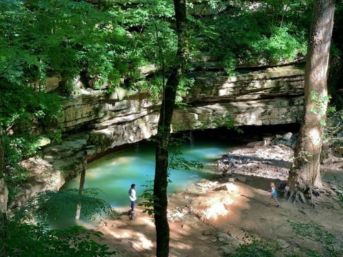 Two people explore a serene, green forest with a rocky overhang and a turquoise pool of water below.