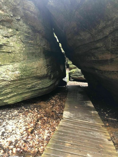 A narrow pathway made of wooden planks between two large rock formations in a forested area.